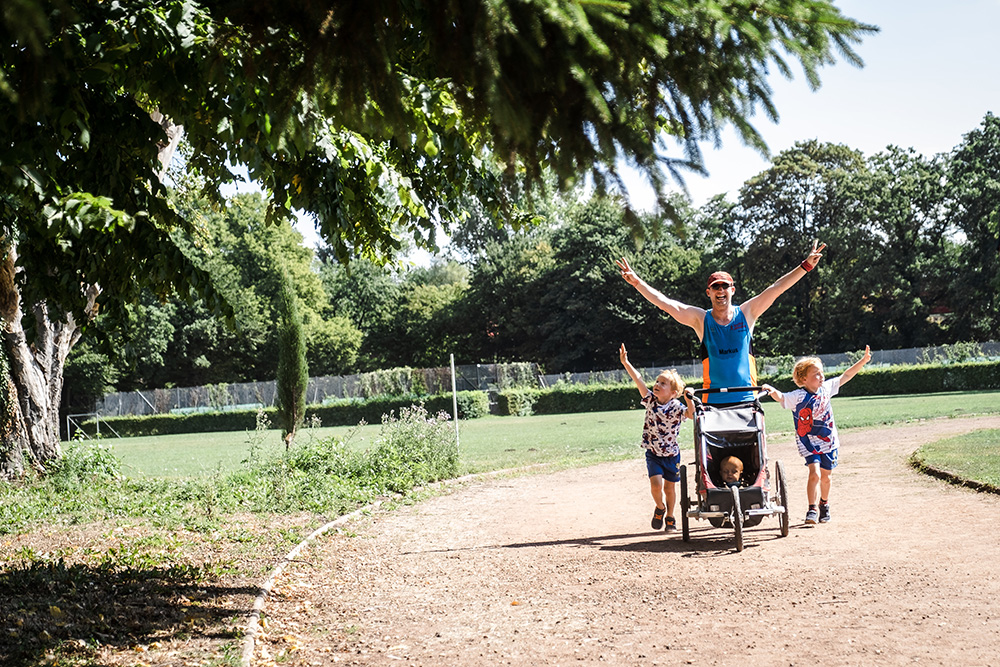 Foto Muko-Spendenlauf Hannover: ein Vater mit zwei Söhnen und einem Kind in einer Laufkarre, alle heben die Hände hoch