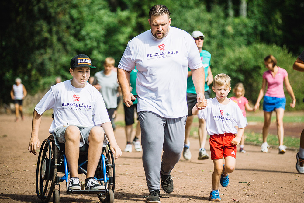 Foto vom Muko-Spendenlauf: ein Junge im Rolli, ein Mann und ein kleiner Junge auf der Laufbahn. Im Hintergrund weitere Läuferinnen und Läufer