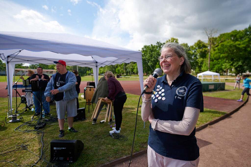 Foto vom 17. Muko-Spendenlauf 2024: Eien Frau steht auf der Laufbahn und hält ein Mikro in der Hand, im Hintergrund baut eine Band auf