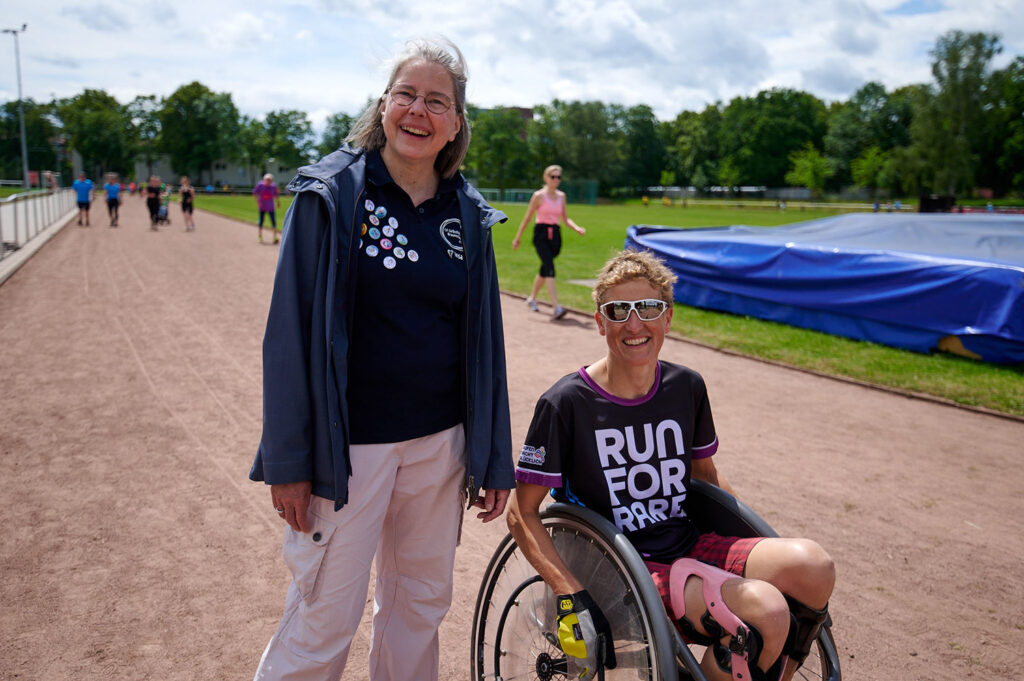 Foto vom 17. Muko-Spendenlauf 2024: Zwei Frauen auf der Laufstrekce, im Hintergrund Läuferinnen und läufer - Insa Krey, stehend, Organisatorin des Muko-Spendenlaufs und Tina Deeken im Rollstuhl, Schirmherrin des Laufs