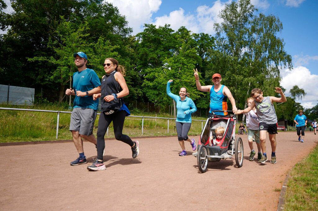 Foto vom 17. Muko-Spendenlauf 2024: Ein Paar läuft auf der Strecke, dahinter eine Familie mit Laufbuggy und Kindern - die Familie hebt die Hand und winkt