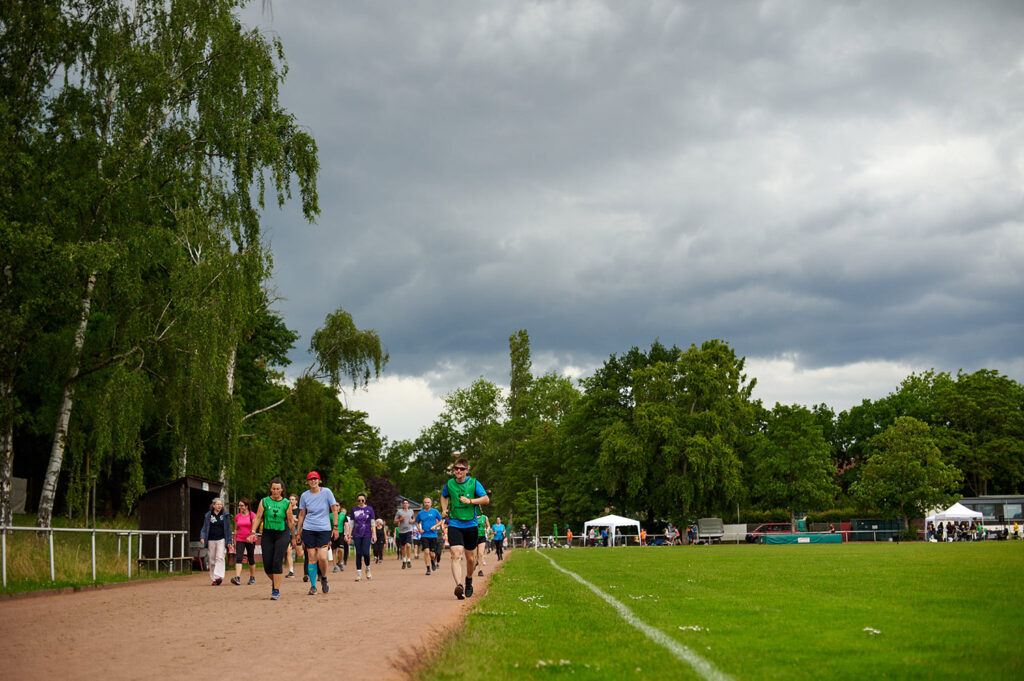 Foto vom 17. Muko-Spendenlauf 2024: Blick auf die Laufbahn und den Sportplatz, viele Läuferinnen und Läufer laufen oder gehen, darüber ist ein Himmel mit dunklen Woken
