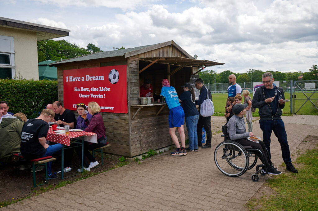 Foto vom 17. Muko-Spendenlauf 2024: Eine Holzbude vor der Vereinsgaststätte, davor und daneben sind verschiedene Personen zu sehen, einige Essen Wurst
