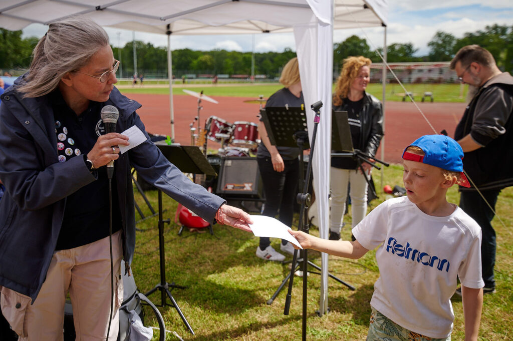 Foto vom 17. Muko-Spendenlauf 2024: Verlosung, eine Frau reicht einem Jungen eine Umschlag, dahinter Mitglieder der Band