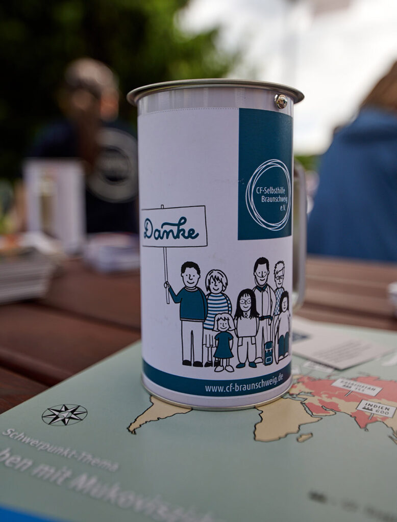Foto vom 17. Muko-Spendenlauf 2024: Spendendose steht auf einer Zeitschrift, auf der Spendendose sind verschiedene Personen gezeichnet, eine Person hält ein Schild mit "Danke" in die Luft, darüber das Logo der CF-Selbsthlife Braunschweig e.V.